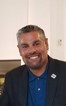 A photo of a male in a blue shirt and black suit jacket smiling