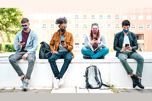 four students sitting on campus wall in front of brick building. all are wearing masks and looking at their phones. backpacks sitting nearby