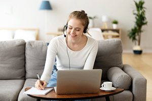smiling woman with headphones sitting on couch at laptop and writing on notepad