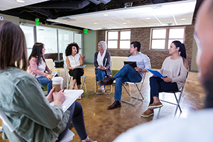 Group of people sitting on folding chairs arranged in a circle while they all hold note pads.