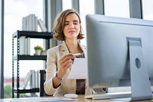 Female professional looking at a paper while sitting in front of a computer monitor at an office desk.