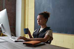 Female of color working on a computer while sitting at a desk in the front of a classroom.