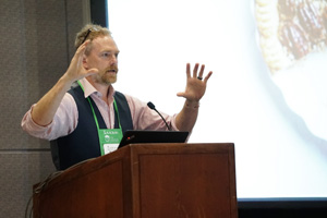 male speaker in a pink dress shirt and grey vest gestures with his hands from behind a podium