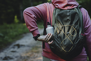 person wearing a red sweatshirt grabs their water bottle from their green backpack while walking on a wooded trail