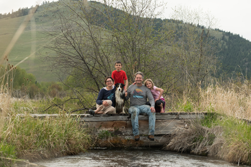 family of four: man, woman, two children under 10, with dog, pose on a rustic wooden bridge over a creek, mountain the background