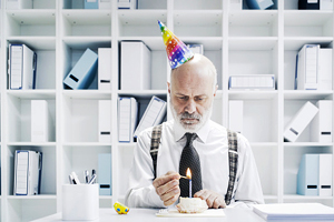 sad man at desk with party hat on lighting candle on cupcake