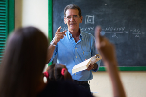 teacher wearing a blue collared shirt points to a student who is raising their hand