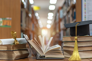 Open book, laptop, and diploma on table in foreground with library in background.