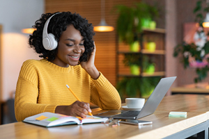 smiling black student with headphones and laptop, writing in textbook