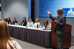 Male speaker at the front of a room with seven panelists seated at a table next to him