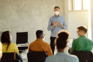 professor in mask teaches seated students in masks in college classroom