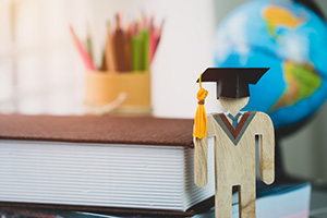wooden figurine wearing a graduation cap standing in front of a book, pencil container, and globe 