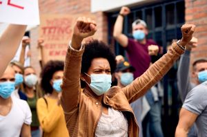 African American woman wearing protective face mask while protesting with arms raised.