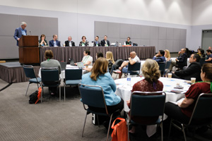event space with people sitting at circular tables as they listen to a group of panelists