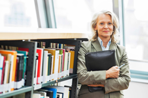 older female dressed in a suit leans against a book case while holding a portfolio book