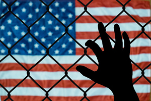 The stars and stripes hang in the background with a silhouette of a hand clinging to a fence in the foreground