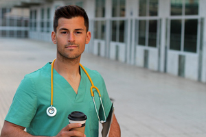 a male in turquoise scrubs with a stethoscope around his neck stands outside with a coffee 