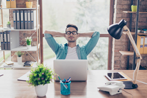 male in glasses stretches his back as he sits at a desk with a laptop, papers, phone, and tablet visible