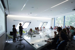 several figures sit around a large white table while two figures stand at the front of the room