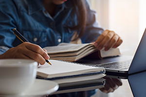 person in a blue flannel takes notes while reading a book and looking at their laptop