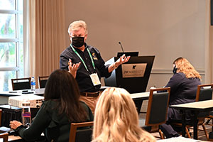 photo of Kevin Pollock wearing a black collared shirt and black face mask while speaking from the front of a room