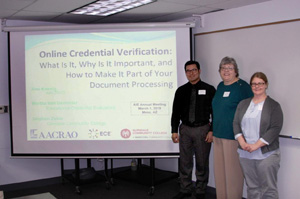 Three people pose for a photo beside a projector screen with the title "Online Credential Verification" visible