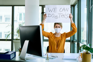 female wearing a face mask while sitting at an office desk holds up a sign that reads "we can do this"