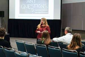 Female dressed in red top stands in front of an audience with a projector screen displaying "Any Questions" behind her