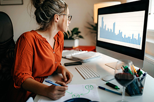 female in orange blouse writes on paper while looking at graphs on her computer monitor