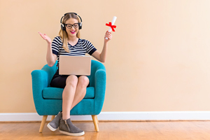 female student sitting on a blue chair celebrates graduating while looking at her laptop