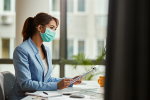 professional woman with ponytail in blue suit wearing mask reading a document at a desk