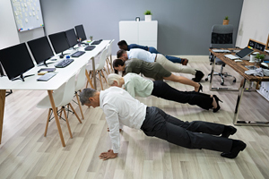 line of 5 people in business attire perform pushups in an office