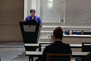 female wearing a blue blouse speaks from behind a podium