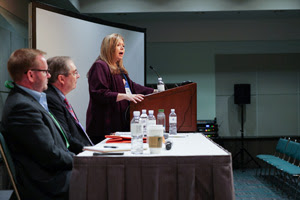 female standing behind a podium with two males seated at a table next to her