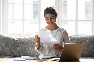 female smiling at a piece of paper while sitting on a couch with her laptop in front of her resting on a coffee table