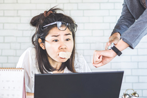 female with glasses askew and donut in mouth appears stressed looking at computer while boss points at watch