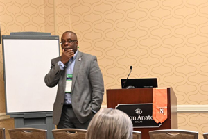 male of color dressed in a suit stands in the front of a room