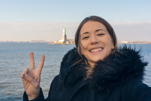 female in a fluffy coat poses for a selfie with the Statute of Liberty in the background 