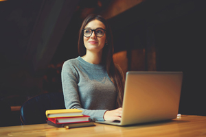 young student smiling out the window, seated at a table with books and laptop