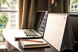 laptop sitting on a desk near a window with a calendar and notepad next to the laptop