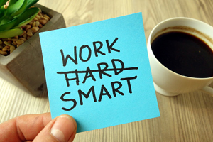 hand holding post-it note reading "work smart" with the word "hard" crossed out above a table holding a cup of coffee and a cactus
