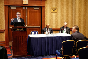 male speaks from behind a podium while 2 other males sit at a panelist table to the speaker's left 
