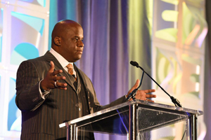 male in a suit with outstretched arms speaks from behind a podium