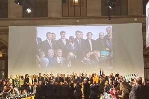 Audience in a large event space all facing a projector screen that has a group of professionals displayed on it.