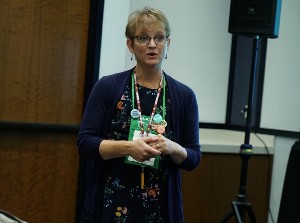 Female wearing a blue blouse as she speaks from the front of an event space.