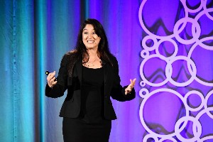 Female presenter wearing a black jacket as she speaks on a stage with a purple and blue lit backdrop.