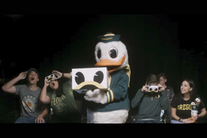Duck mascot wearing green as they stand next to a group of students making faces of amazement.