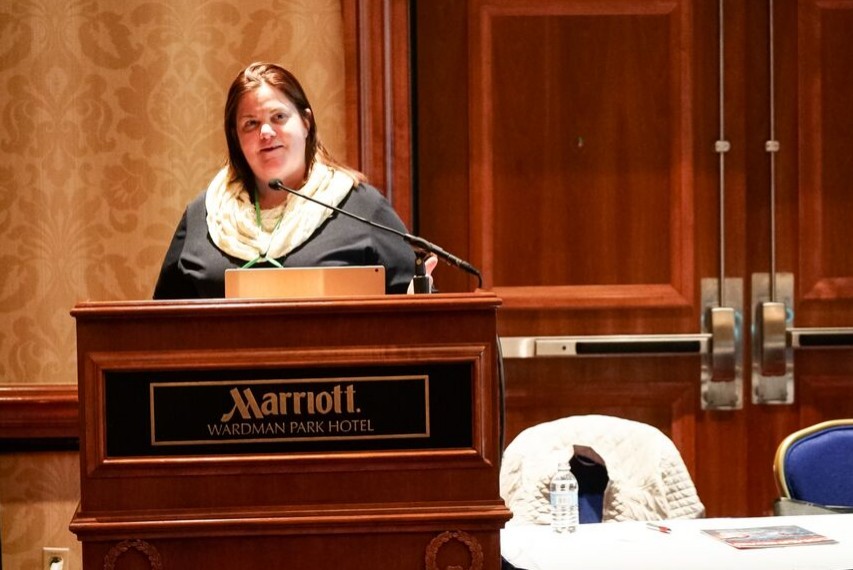 Female wearing a black top as she speaks from behind a wooden podium that says "Marriott Wardman Park Hotel."