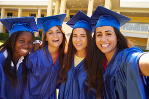 Four females wearing blue graduation caps and gowns smiling as they take a group selfie together.