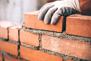 Gloved hand building a wall of red brick and mortar.